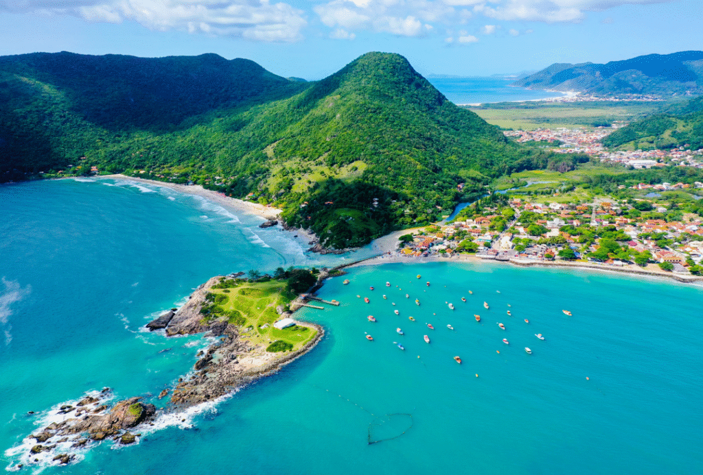 Aerial view of a lush green coastal landscape in Florianópolis, Brazil, featuring turquoise bays, sandy beaches, forest-covered hills, and a small seaside town under a bright blue winter sky