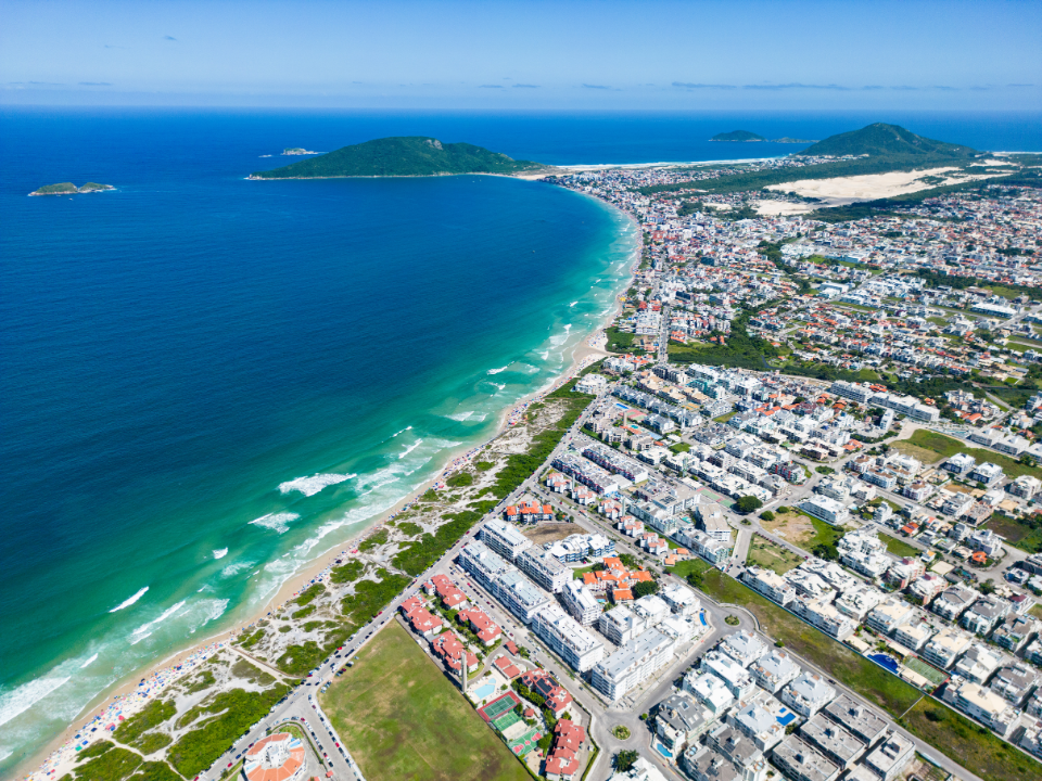 Aerial view of Florianópolis, Brazil, showing beachfront neighborhoods, residential buildings, turquoise ocean waters, and coastal urban development along the island's shoreline