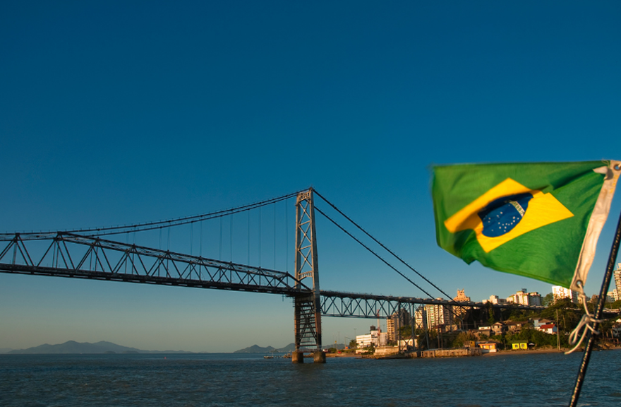 Brazilian flag displayed beside a large suspension bridge over water at sunset, with the bridge and coastal landscape visible in the background