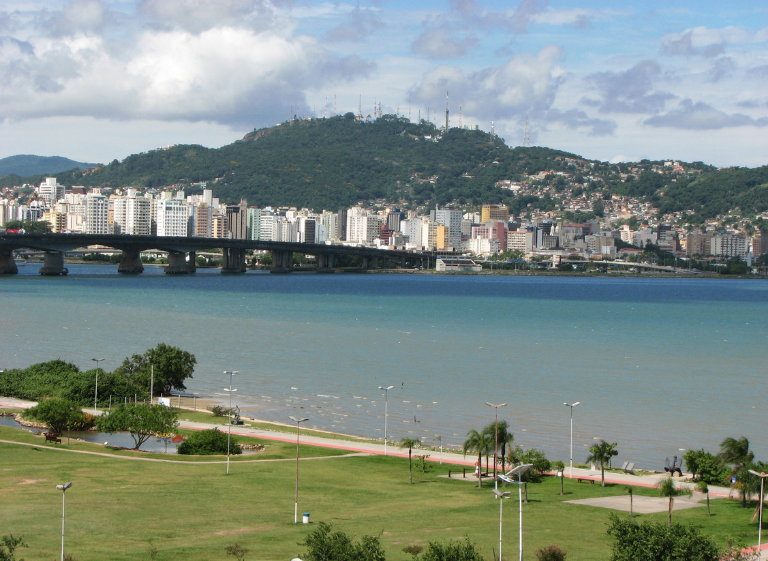 Panoramic view of Florianópolis, Brazil, featuring a calm bay, waterfront park, modern city skyline, a long bridge, and lush green hills rising in the background.