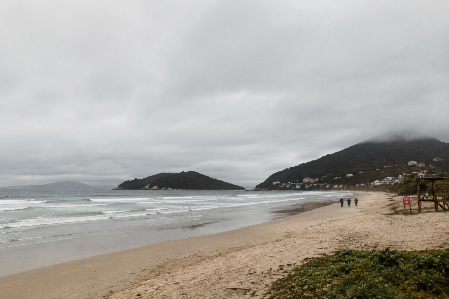 Quiet Florianópolis beach on an overcast winter day, with grey skies, gentle waves, wet sand, and misty hills in the background