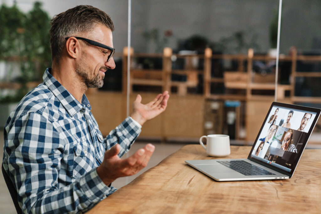 A man wearing glasses gestures during a video conference call on a laptop while sitting at a wooden table in an office