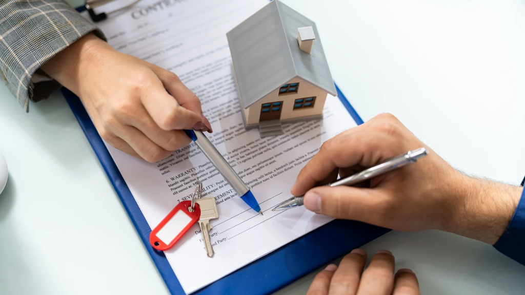 Hands signing a property purchase agreement with a miniature house model, house keys, and a pen on real estate documents, representing property prices and buying costs.