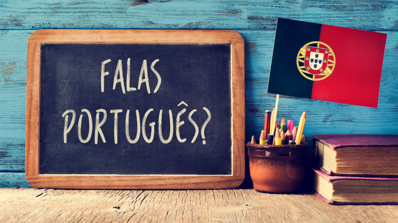 Chalkboard displaying “Falas Português?” in Portuguese, alongside the Portuguese flag, a pot of coloured pencils, and stacked books on a rustic blue wooden table.