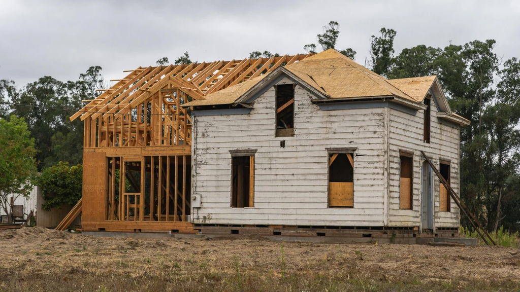 House under renovation with exposed wooden roof trusses, unfinished timber framing, and weathered white exterior siding at a residential construction site surrounded by trees